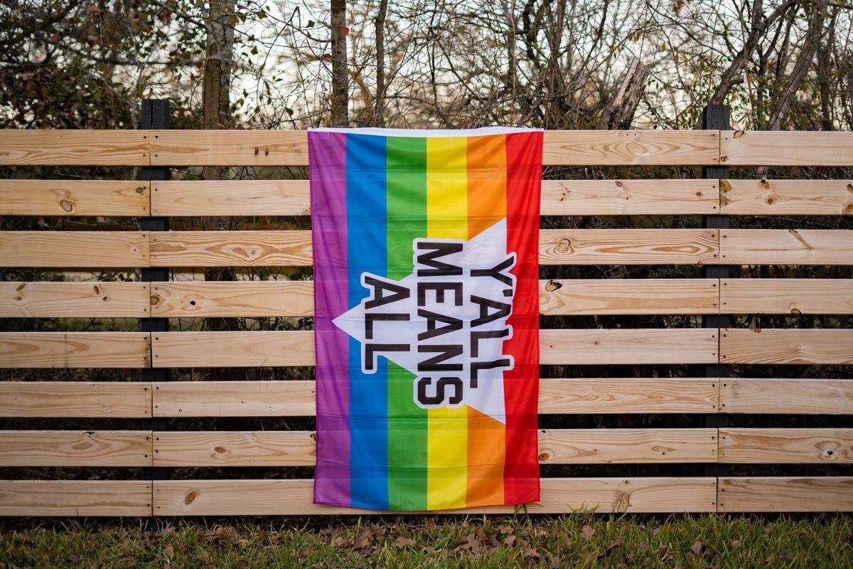 Y'all Means All rainbow flag displayed on a fence outdoors